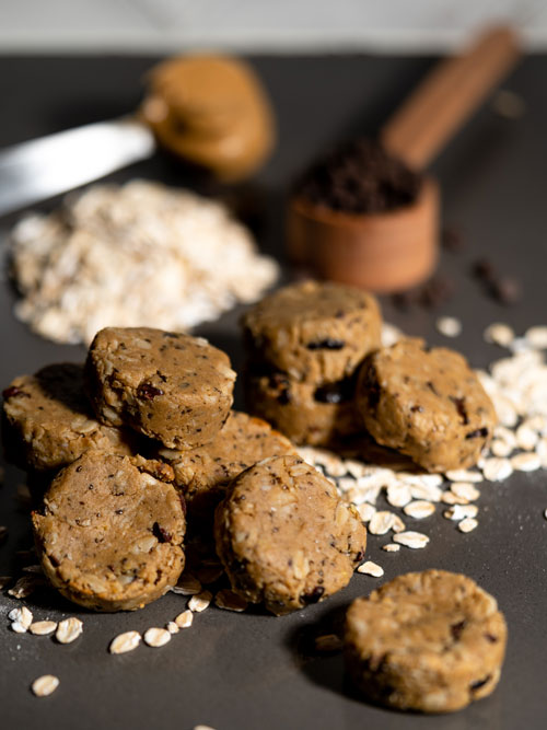 stack of 9 energy bites with oats and cocoa nibs on top of oats with peanut butter and cocoa nibs in the background.