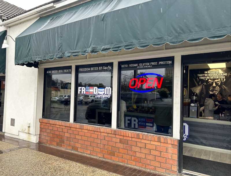 Red, white, and blue sign in a businesses setting that says 'Freedom Bowls Superfoods' with a lit up red open sign hanging in the window and a green overhang on a brick building. 