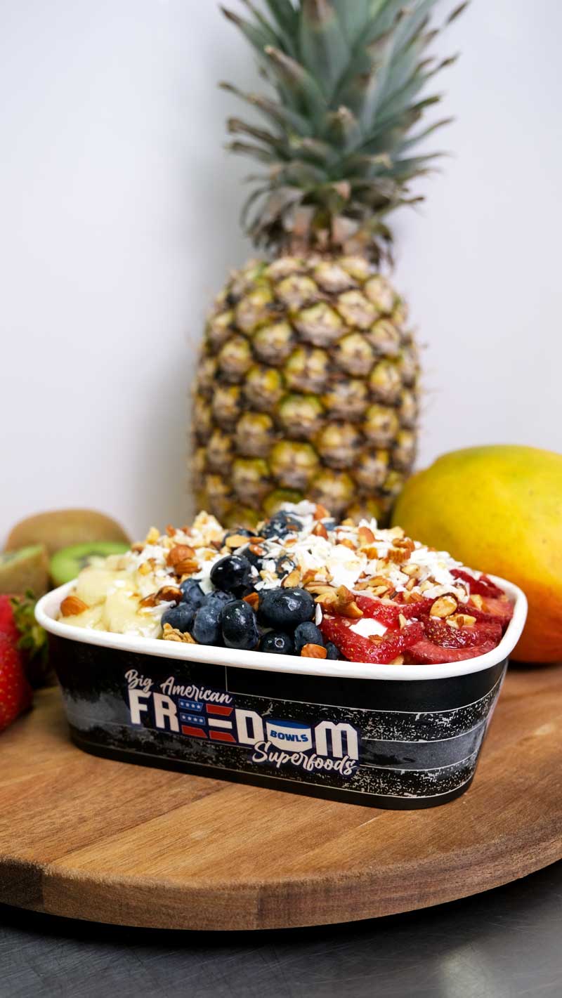 Bright colored fruit bowl with chopped strawberries, blueberries, sliced bananas, chopped almonds and shredded coconut granola in a navy blue to-go container that says 'Big American Freedom Bowls Superfoods'.