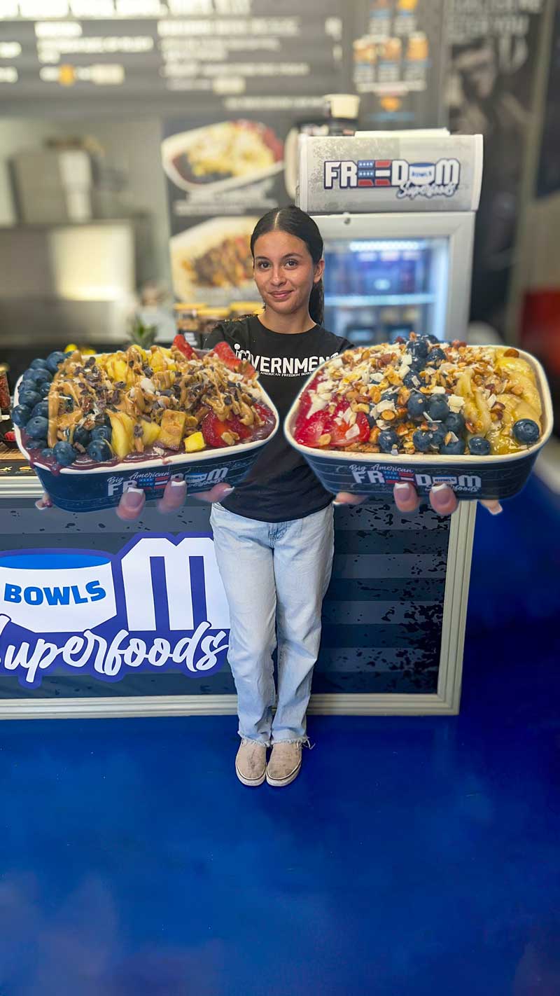 Bright colored fruit bowl with chopped strawberries, blueberries, sliced bananas, chopped almonds and shredded coconut granola in a navy blue to-go container that says 'Big American Freedom Bowls Superfoods'.